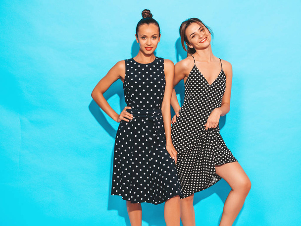 Two young beautiful smiling hipster girls in trendy summer polka-dot dresses.Sexy carefree women posing near blue wall.Having fun and hugging. Models shows good relationship. Female with red lips - Photo, image
