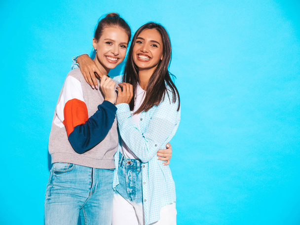Dos jóvenes hermosas y sonrientes chicas hipster rubias en ropa de moda de verano colorida camiseta. Sexy mujeres despreocupadas posando cerca de la pared azul. Modelos positivos divirtiéndose
 - Foto, imagen
