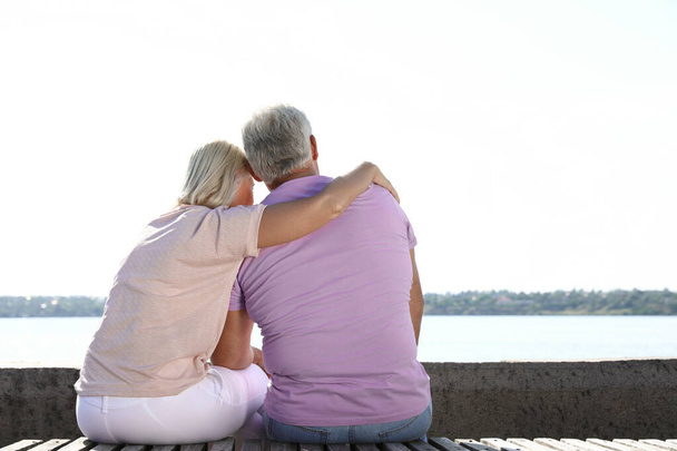 Affectionate senior couple sitting near river together on sunny day. Space for text - 写真・画像
