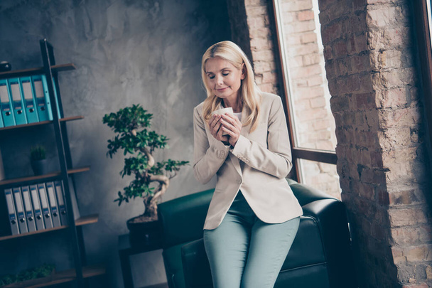 Portrait of charming peaceful dreamy middle aged woman hold cup mug with coffee smell enjoy rest before hard start-up workshop seminar wear beige blazer sit on couch in enterprise office - Foto, afbeelding