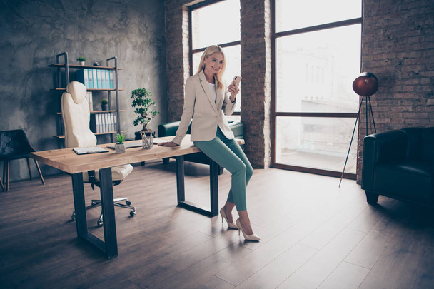 Full size photo of confident focused middle aged leader business woman sit on wooden table in office use her smartphone chat with her clients workstation partners wear high-heels - Photo, Image