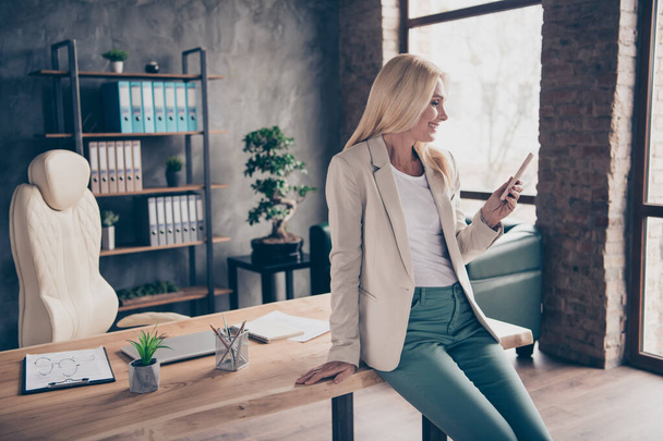 Portrait of positive confident cool blonde hair middle aged businesswoman use her cellphone type messages communicate chatting with work partners investors sit on wooden table in office workplace - Φωτογραφία, εικόνα