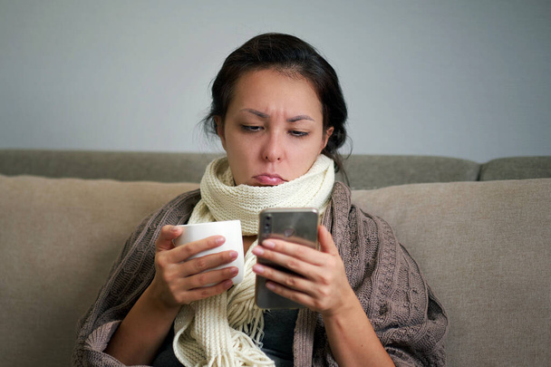 Ill upset woman blowing running nose got flu caught cold sneezing in tissue sit on sofa, sick girlsore throat, gray with a scarf throat and drinks aspirin at home, hay fever concept - Photo, Image