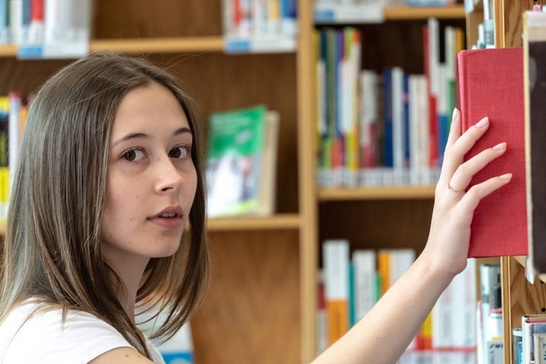 Fiatal diáklány választ és keres egy könyvet a könyvespolcon az iskolai könyvtárban - Fotó, kép