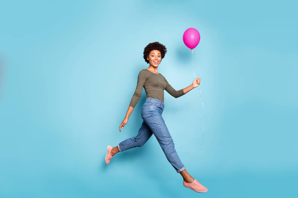 Full length photo of dark skin pretty wavy lady jumping high walking down street moving with help of air balloon wear casual outfit isolated pastel blue color background - Фото, изображение