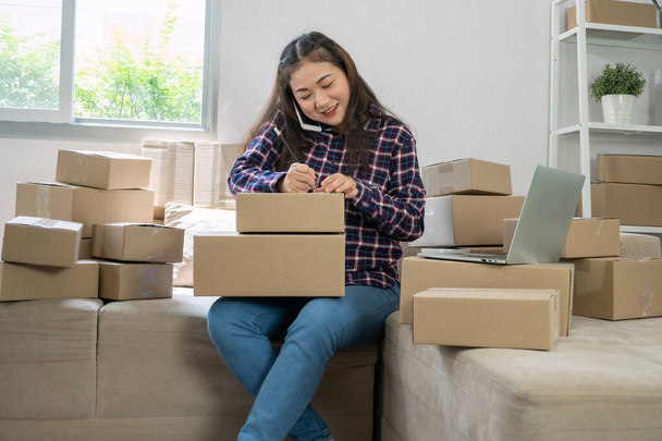 Young entrepreneur, sitting on the sofa and working at home. Receiving orders from customers by phone and writing customer address deliver shipment online sales. Concept of small business owner. - Photo, Image