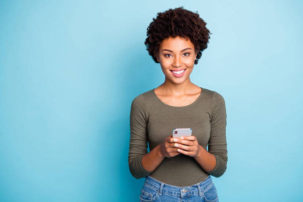 Portrait of positive cheerful dark skin girl use hold her smartphone enjoy blog posts comments wear green outfit isolated over blue color background - Foto, afbeelding