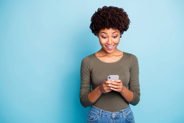 Portrait of amazed funky mulatto girl use her smartphone read feednews got like from favorite blogger feedback scream wow omg wear green stylish pullover isolated over blue color background - Photo, Image