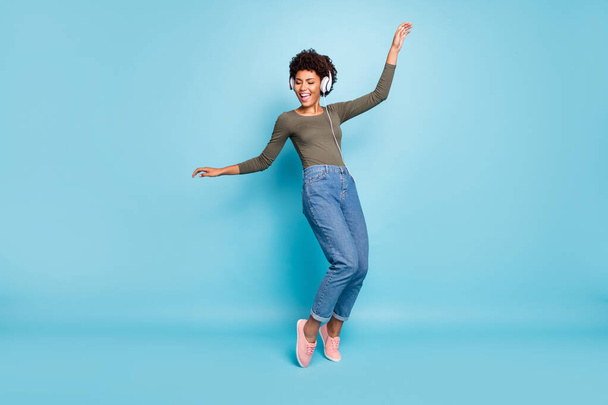 Full body photo of cheerful funny mulatto girl have free time listen music on her headset dance sing enjoy wear stylish green pullover denim jeans sneakers isolated over blue color background - Fotografie, Obrázek