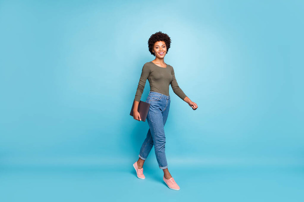 Teljes méretű fotó pozitív magabiztos független hűvös sötét bőr lány szabadúszó megy sétálni a munkát a cég tartsa neki netbook viselni alkalmi stílusú ruhák rózsaszín tornacipő elszigetelt kék színű háttér - Fotó, kép