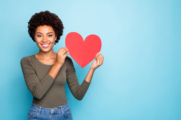 Portrait of positive dreamy dark skin girl hold big red paper card valentine heart for 14-february holidays enjoy date with sweetheart wear green sweater denim jeans isolated blue color background - Fotoğraf, Görsel