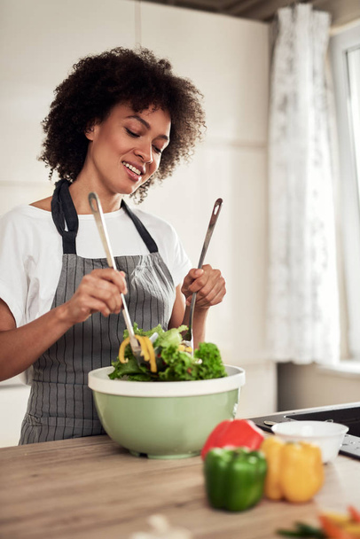 Attractive mixed race woman in apron mixing vegetables in bowl while standing in kitchen at home. - Foto, Bild