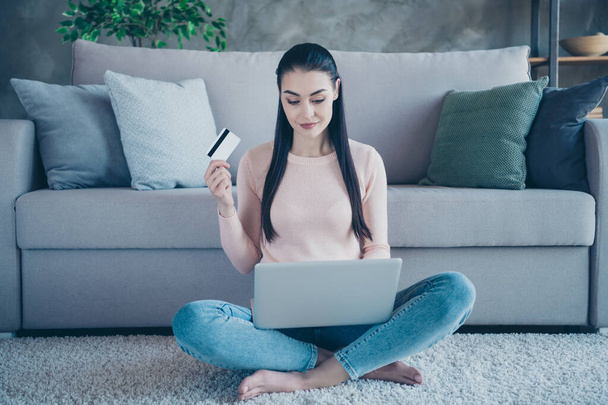 Photo of addicted lady holding notebook on knees going to pay online with help of credit card sitting floor on comfy carpet wearing pullover and jeans in apartment - Фото, зображення