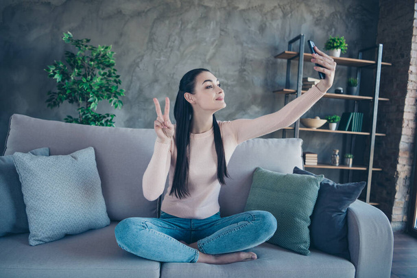 Φωτογραφία της όμορφης κυρίας με τηλέφωνο στα χέρια κάνοντας selfies δείχνει σύμβολο v-σημάδι κάθεται άνετα στον καναπέ φορώντας πουλόβερ και τζιν homey στο διαμέρισμα - Φωτογραφία, εικόνα