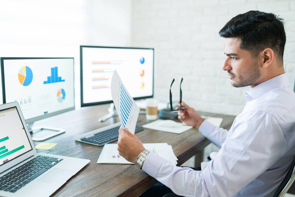 Aantrekkelijke jonge mannelijke sales professional analyseren grafiek terwijl het vasthouden van papier aan het bureau - Foto, afbeelding