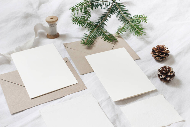 Christmas table, festive styled composition. Christmas greeting cards, envelopes mock-ups. Silk ribbon, pine cones and fir tree branch on white linen background. Top view. Festive craft, stationery. - Fotó, kép