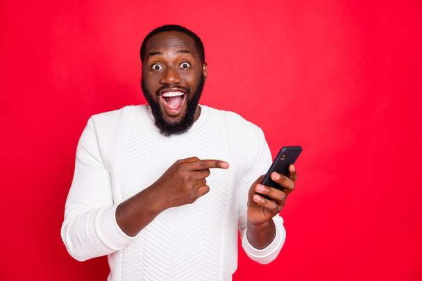 Photo of funny dark skin man holding telephone advertising new model smart phone indicating finger advice low price wear white knitted sweater isolated red background - Fotoğraf, Görsel