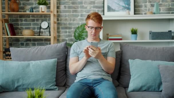 Cheerful teenager using smartphone relaxing on sofa at home enjoying gadget - Footage, Video