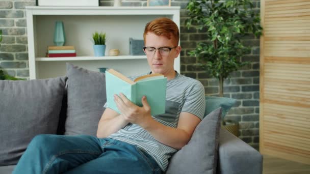 Handsome guy in glasses reading book and smiling sitting on sofa in apartment - Felvétel, videó