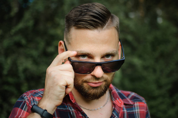 Outdoors portrait of young bearded man with sunglasses looking at the camera in the park on trees background - Photo, Image