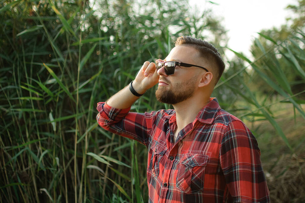 На відкритому повітрі портрет молодого бородатого чоловіка з сонцезахисними окулярами, який дивиться на камеру в парку на фоні дерев
 - Фото, зображення