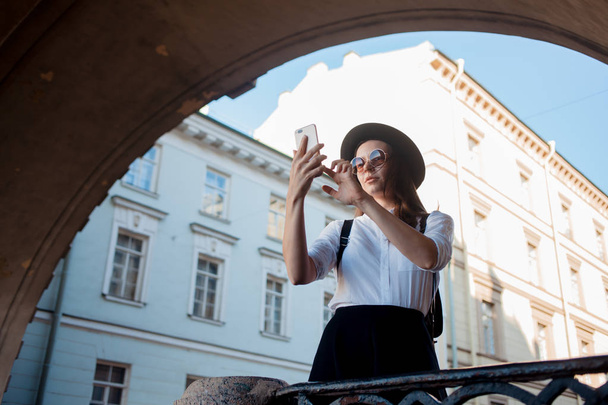 Tourist in the city takes a photo on smartphone. A young woman in a black hat and a white shirt, - Foto, immagini