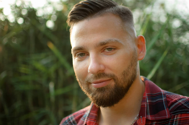 Brutal bearded young man in red shirt is standing outdoors on green stems background. Hipster style concept. - Photo, Image
