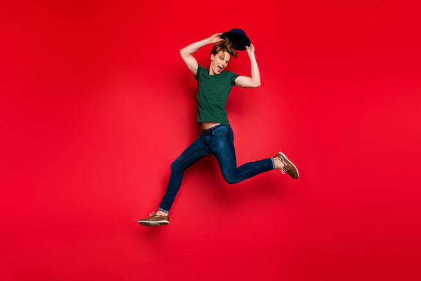 Full length profile side photo of cheerful crazy guy jump run have fun hold his blue headwear wear casual style green t-shirt sneakers isolated over red color background - Fotó, kép