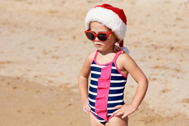 Little girl child in a swimsuit and Santa hat walks on a tropical beach on the ocean. The concept of New Year and Christmas holidays. Tourism and rest - Foto, imagen