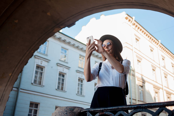 Tourist in the city takes a photo on smartphone. Young woman in black hat and white shirt. - Zdjęcie, obraz
