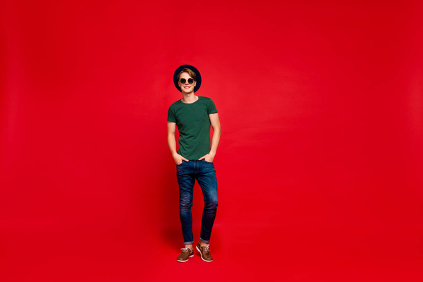 Full length photo of positive cheerful guy in blue hat real wealthy rich modest millionaire have fun on summer holidays put hands in denim jeans outfit isolated over red color background - Photo, Image