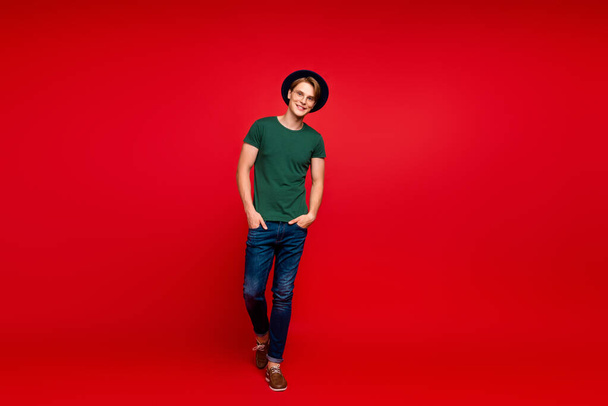 Full size photo of charming rich wealthy modest guy put hands in pockets go walk on summer weekends wear blue headwear glasses green t-shirt denim jeans isolated over red color background - Foto, imagen
