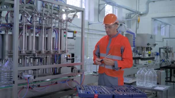 fabrieksservice, technicus in helm en werkkleding reparaties fabrieksuitrusting met speciaal gereedschap in de buurt van transportband voor het bottelen van mineraalwater in plastic flessen in de fabriek - Video