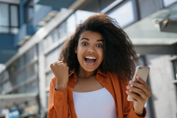 Nadšený Afroameričanka vyhrát online loterie, slavnostní úspěch. Happy emotivní dívka pomocí smartphone s mobilní aplikací pro on-line nakupování s nízkou cenou, v hotovosti zpět na černý pátek - Fotografie, Obrázek