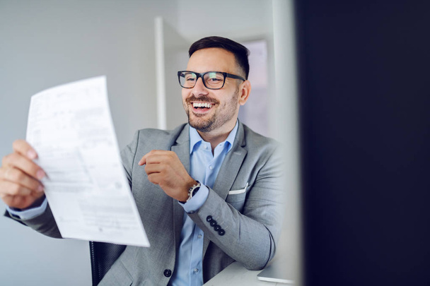 Усміхнений працьовитий кавказький менеджер у сірому костюмі сидить у сучасному офісі та читає звіт
. - Фото, зображення