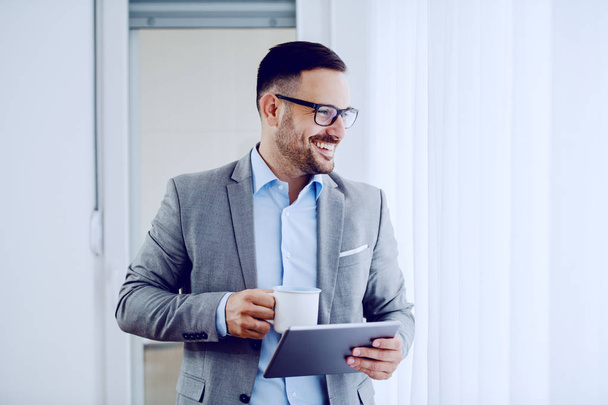 Όμορφος μοντέρνος χαμογελαστός άντρας με κοστούμι και με γυαλιά που πίνει καφέ, κρατάει tablet και κοιτάζει από το παράθυρο. Εσωτερικό. - Φωτογραφία, εικόνα