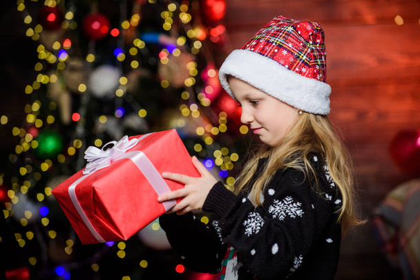 Girl cute child hold wrapped gift near christmas tree. Winter shopping. Holiday party that actually sounds fun. Merry christmas. Kid adorable wear santa hat celebrate christmas. Buy christmas gift - Photo, Image