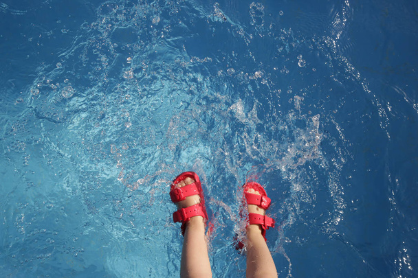 Дитина сидить біля краю басейну і розбризкує воду в басейні, дитячі ноги з червоними гумовими сандалі у воді, концепція літньої релаксації сімейного відпочинку
 - Фото, зображення