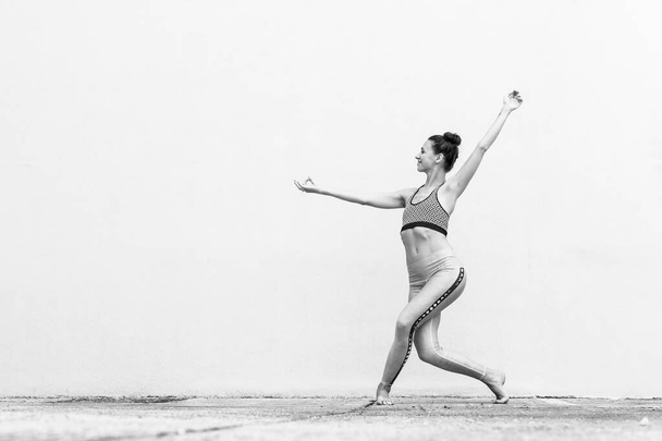 Впишеться спортивна активна дівчина в модний спортивний одяг, займаючись фітнесом йоги перед сірою стіною, спортом на відкритому повітрі, міським стилем. Чорно-біле фото
. - Фото, зображення