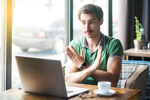 Young serious businessman in green t-shirt looking at laptop screen during video call and showing closed sign with hands while sitting in cafe, business and freelancing concept - Photo, image