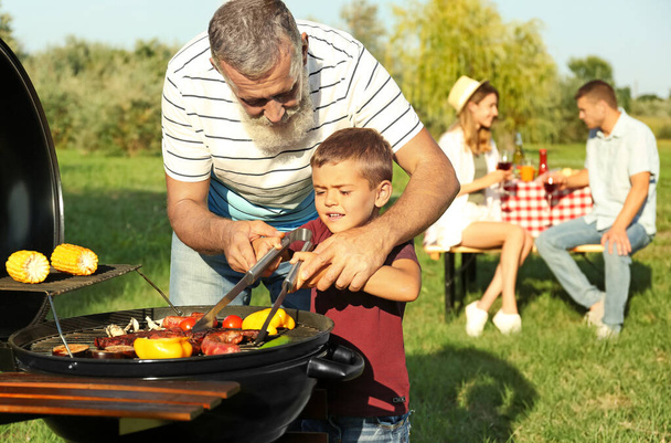 Grandfather with little boy cooking food on barbecue grill and their family in park - Fotó, kép