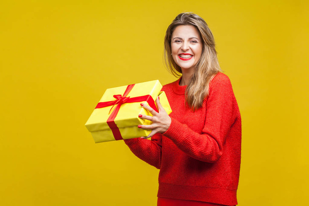 charming blonde woman with red lipstick in bright casual sweater holding christmas wrapped gift box and smiling at camera on yellow background - Фото, изображение
