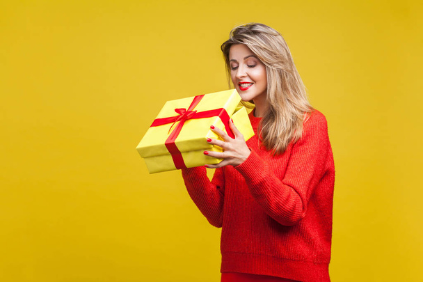 curious beautiful blonde woman with red lipstick in bright casual sweater looking christmas inside gift box while posing on yellow background - Foto, Imagen