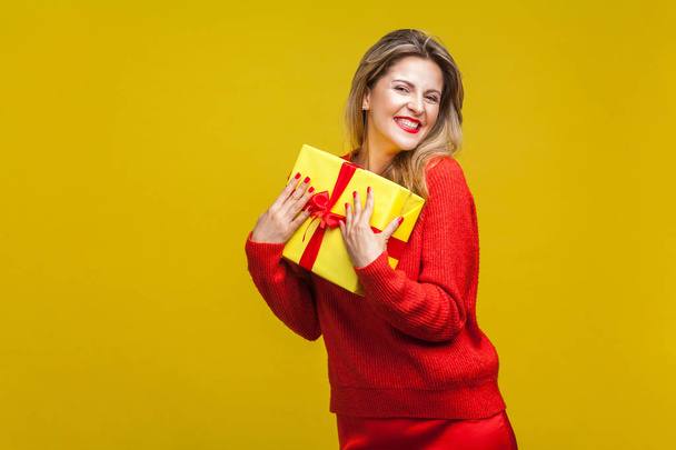 happy woman with red lipstick in bright casual sweater holding wrapped gift box and smiling at camera on yellow background, birthday surprise concept  - Foto, Bild