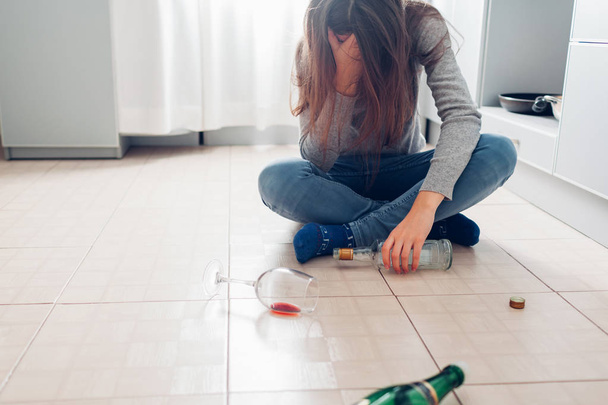 Female alcohol addiction. Young woman sitting on kitchen floor after party and holding bottle. Hangover - 写真・画像