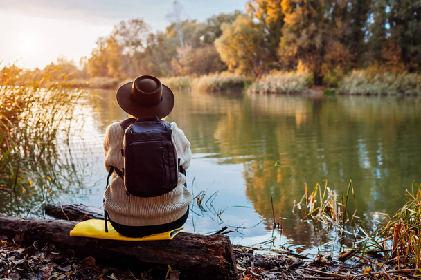 Tourist with backpack sitting on river bank at sunset. Middle-aged woman admiring autumn nature - Foto, Imagen