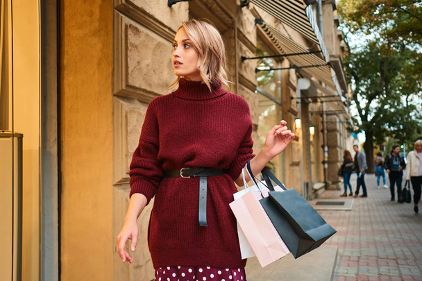Mooi stijlvol blond meisje in gebreide trui met boodschappentassen dromerig wandelen op straat - Foto, afbeelding