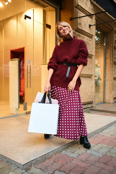 Hermosa chica rubia sonriente con estilo con bolsas de compras mirando alegremente en la cámara después de comprar al aire libre
 - Foto, Imagen