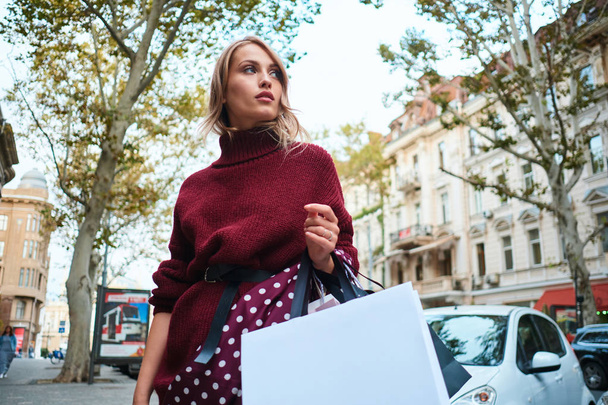 Πανέμορφο κομψό ξανθό κορίτσι με τσάντες ψώνια προσεκτικά κοιτάζοντας μακριά στο δρόμο της πόλης - Φωτογραφία, εικόνα