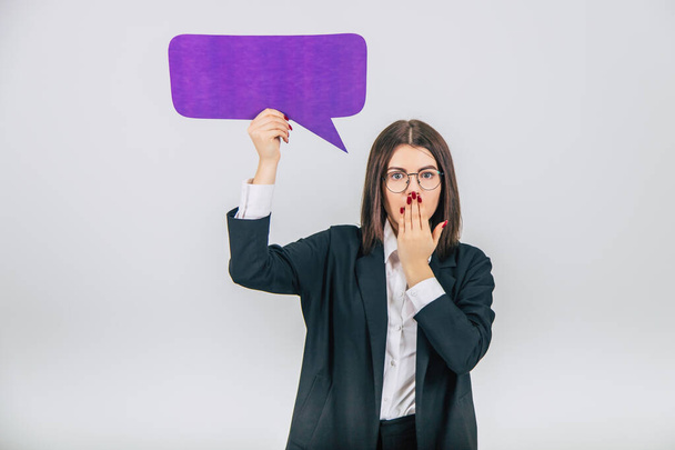 Portrait of an excited young business woman holding empty square violet speech bubble, covering her mouth with hand, surprised face. - Photo, image